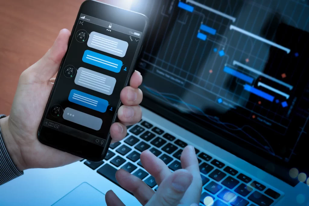 Hand using chatbot app on smartphone in front of a laptop with graphs